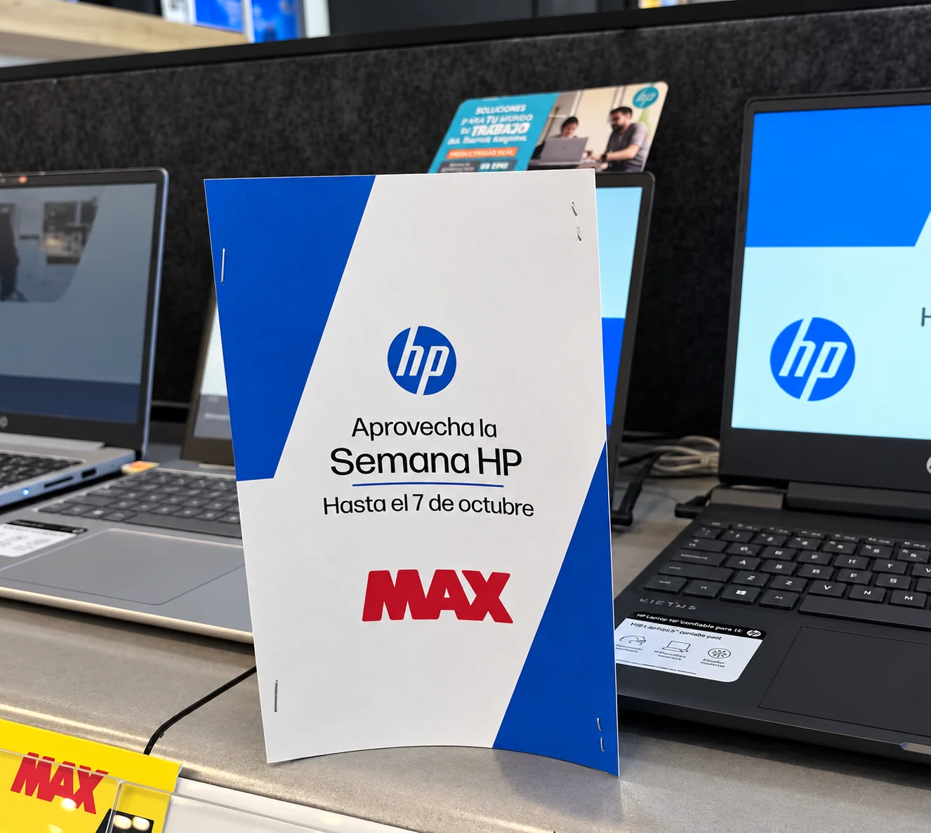 Afiche de la Semana HP sobre una mesa de exhibición de laptops en una tienda MAX